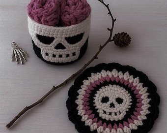 May include: A crochet skull-shaped container with a purple brain inside, a matching coaster, and a skeleton hand. The container is black and white, and the coaster has a skull design in the center.