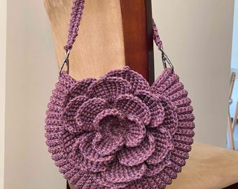 May include: A handcrafted, round, purple crochet bag with a flower design. The bag features a detailed, layered petal pattern and a matching strap with silver-tone hardware. The bag is made of a soft, textured yarn.