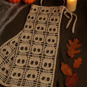 May include: A beige crochet apron featuring a repeating skull pattern, perfect for Halloween. The apron has a long, slender design with a neck strap and ties. The image includes pumpkins, leaves, and a lit candle, creating a festive autumn scene.