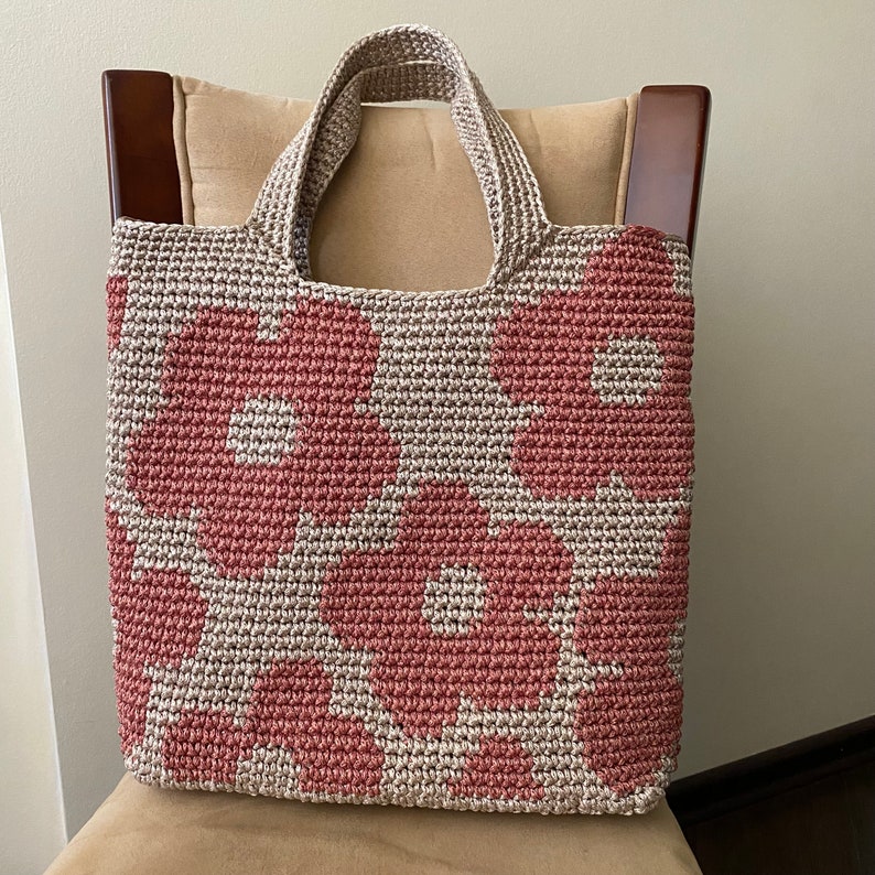 May include: A handmade tote bag with a floral pattern. The bag is crocheted with a light beige and dusty rose color scheme. The handles are also crocheted. The bag is sitting on a beige chair.