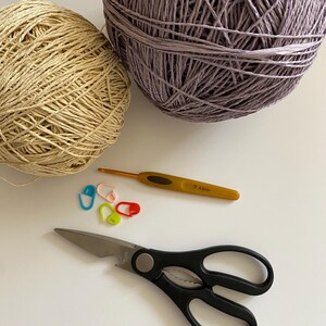 May include: Overhead shot of crochet supplies. Two yarn balls, one beige and one purple, are next to a yellow crochet hook, stitch markers, and black scissors. The hook is labeled "7 4.5mm".