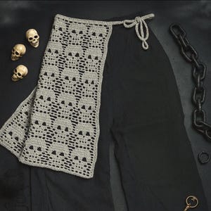 May include: A gray crochet scarf featuring a skull pattern, draped over black fabric. Three small skull figurines are visible to the left. A black chain and other gothic-themed accessories are also present.