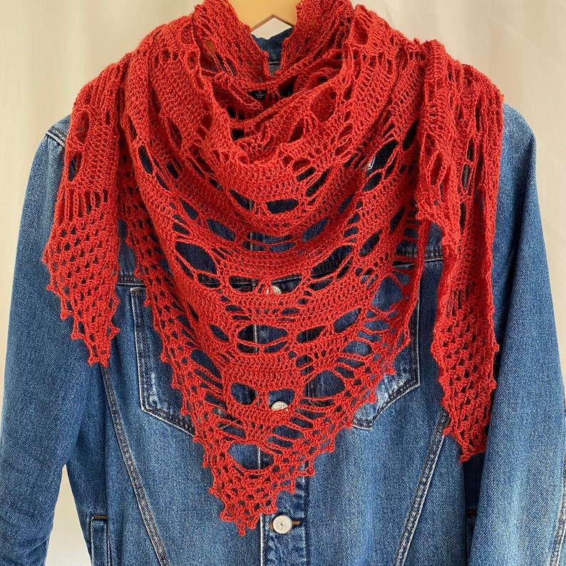 Puede incluir: Un chal de ganchillo rojo con un dise&ntilde;o de encaje abierto. El chal est&aacute; drapeado sobre una chaqueta vaquera.