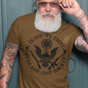 May include: A brown t-shirt with a black graphic of an eagle and crossed rifles. The graphic says "In God We Trust Guns Are Just Backup".