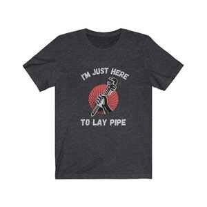 May include: Dark heather grey t-shirt with a red and white graphic of a hand holding a wrench. The text "I'm just here to lay pipe" is printed in white on the shirt.