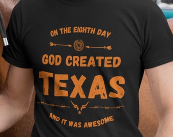 May include: Black t-shirt with orange text that reads "On the eighth day God created Texas and it was awesome". The text is surrounded by a star, arrows, and barbed wire.