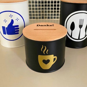 Könnte beinhalten: Drei Metall-Sparschweine mit Holzdeckeln. Die erste Spardose ist weiß mit einem blauen Daumen-hoch-Symbol und Sternen. Die zweite Spardose ist schwarz mit einem goldenen Kaffeetassen-Symbol und dem Wort "Danke!" auf dem Deckel. Die dritte Spardose ist schwarz mit einem weißen Gabel-, Messer- und Löffel-Symbol.