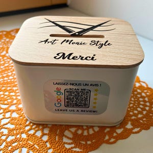 Op de afbeelding: Een witte doos met een houten deksel. Het deksel heeft de tekst "Art Monic Style" en "Merci", een gleuf en een schaargrafiek. Aan de zijkant zit een Google review sticker.