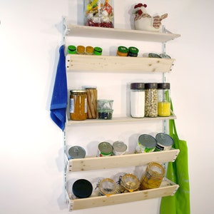 May include: White wall mounted shelving unit with four shelves and a blue towel hanging on the left side. The shelves are filled with various jars and cans of food. A green apron hangs on the right side.
