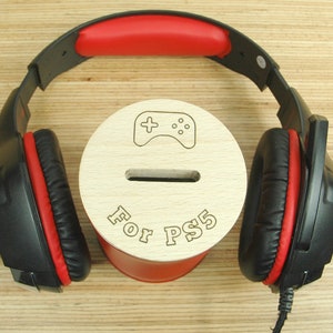 Può includere: Un'auricolare da gioco nero e rosso con microfono posizionato su un salvadanaio di legno con le parole "For PS5" e un simbolo di controller per videogiochi.