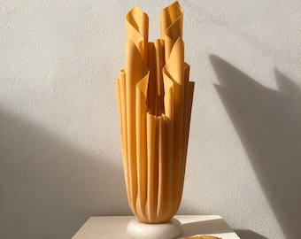 May include: A tall, decorative vase with a unique, folded design in a warm yellow hue. The vase sits on a white, circular base and is displayed on a textured, off-white surface. A single, dried leaf rests nearby, suggesting an autumnal theme.