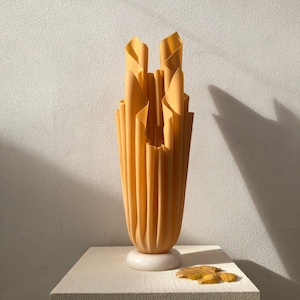 Puede incluir: Un jarrón decorativo alto de color amarillo cálido con un diseño plegado único. El jarrón se apoya sobre una base blanca circular y se exhibe sobre una superficie texturizada blanquecina. Una hoja seca descansa cerca.