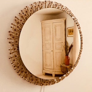 Op de afbeelding: Een ronde spiegel met een decoratief frame van witte bloemen. De spiegel reflecteert een witte kast met twee deuren en een kleine tafel met een vaas met gedroogde bloemen.
