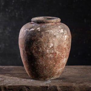 Puede incluir: Un jarrón de cerámica envejecido, de estilo antiguo, con cuerpo ancho y cuello estrecho. El jarrón tiene una superficie moteada y texturizada con tonos marrones, rojos y blancos. Se encuentra sobre una superficie de madera rústica, sobre un fondo oscuro.