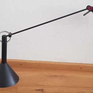 May include: A black desk lamp with a long, adjustable arm and a black, cone-shaped base. The lamp has a red knob on the arm and a white, circular light diffuser.