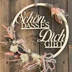 Könnte beinhalten: Ein Holz-Kranz mit dem Text "Schön dass es dich gibt" in weißer Schrift. Der Kranz ist mit getrockneten Blumen in den Farben Rosa, Weiß und Grau verziert.