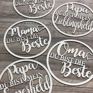 Könnte beinhalten: Fünf runde Holzschilder mit weißer Beschriftung. Die Schilder sagen "Papa du bist der Beste", "Papa du bist mein Lieblingssheld", "Mama du bist die Beste", "Oma du bist die Beste" und "Papa du bist mein Superheld".