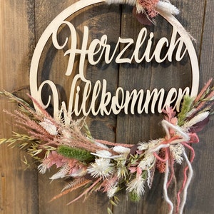 Könnte beinhalten: Ein Holz-Reifen mit den Worten "Herzlich Willkommen" in Schreibschrift. Der Kranz ist mit getrockneten Blumen in den Farben Rosa, Weiß und Beige verziert.