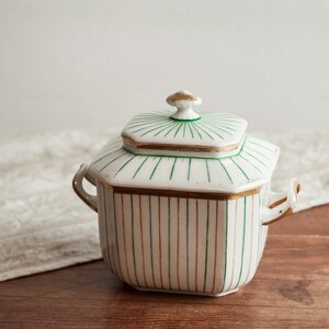 Może przedstawiać: Biała ceramiczna cukiernica z pokrywką, z zielonymi pionowymi paskami i złotymi obramowaniami. Pokrywka ma zielono-złoty wzór. Cukiernica ma dwa małe uchwyty i mały uchwyt na pokrywce. Na drewnianej powierzchni.