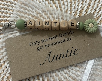 May include: A keychain with wooden letter beads spelling "AUNTIE". The keychain includes a green bead, a flower charm, and a silver clasp. A small card reads "Only the best friends get promoted to Auntie".