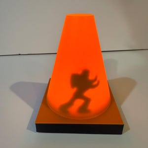May include: An orange cone-shaped lamp with a black base. The lamp casts a shadow of a person. The lamp is on a square orange base. The shadow is of a person in a running pose.