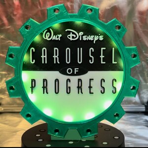 May include: A circular green gear-shaped sign with a black and white center featuring the text Walt Disney's CAROUSEL OF PROGRESS. The sign is illuminated by internal lights and mounted on a black circular base with small white circular accents.