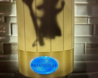 May include: A white night light with a silhouette of Remy from the movie Ratatouille. The night light has a blue button that says "Ratatouille".