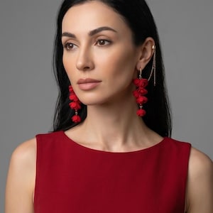 May include: A woman wearing red floral earrings and a matching red dress. The earrings cascade down with multiple flower-shaped clusters. A gold ear cuff with a chain is also visible.
