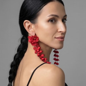 Puede incluir: Pendientes colgantes florales rojos. Los pendientes presentan un grupo de flores en la oreja y elementos florales en cascada. Los pendientes son de color rojo vibrante y los lleva una persona con cabello oscuro.