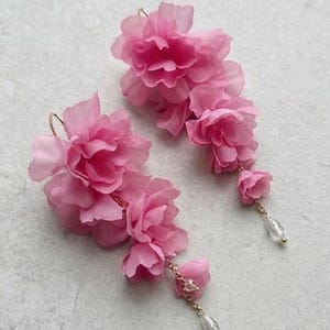 May include: A pair of pink floral earrings. Each earring has multiple layers of pink fabric petals, forming a cascading flower design. Gold-coloured chains hang from the flowers, each holding a clear, teardrop-shaped crystal. The earrings are set against a light background.