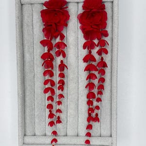 May include: A pair of long, dangling earrings featuring vibrant red fabric flowers at the top. Each earring consists of multiple cascading strands adorned with small, delicate red petal-shaped accents and tiny gold-toned beads, creating a flowing, layered appearance.