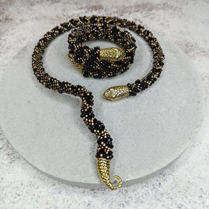 Op de afbeelding: Een zwarte en gouden kralen slangenketting met gouden slangenkop accenten. De ketting is opgerold en ligt op een grijs oppervlak.