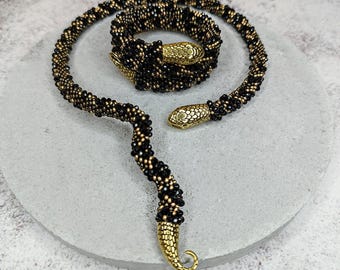 Puede incluir: Un collar de serpiente de cuentas negras y doradas con detalles de cabeza de serpiente dorada. El collar está enrollado y colocado sobre una superficie gris.