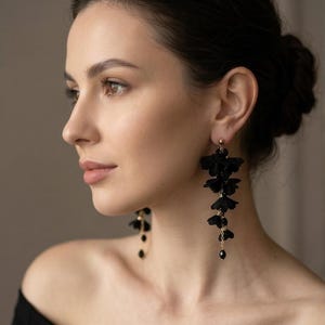 May include: Black floral drop earrings with gold detailing. The earrings have multiple fabric flower shapes, with small black beads at the bottom. The earrings are attached to a gold stud.