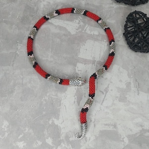 Op de afbeelding: Een rode, zwarte en grijze kralenketting in de vorm van een slang. De ketting heeft een zilveren slangenkoppenhanger.