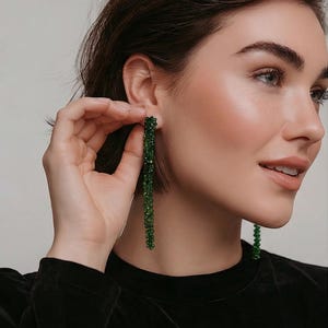 Puede incluir: Una mujer lleva pendientes largos y colgantes de cuentas verdes. Los pendientes están hechos de pequeñas cuentas redondas ensartadas para crear un efecto de cascada. La mujer lleva una blusa negra y tiene el pelo corto castaño.