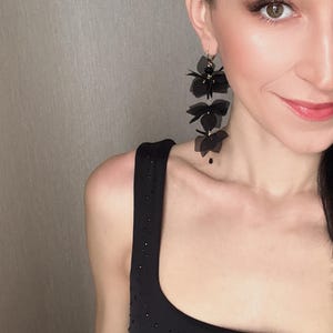 Könnte beinhalten: Schwarze Hängeohrringe mit mehreren Schichten aus schwarzen Stoffblumen. Die Ohrringe werden von einer Person mit dunklen Haaren und Make-up getragen.