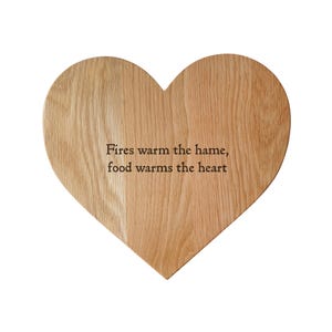 以下が含まれることがあります： 「Fires warm the hame, food warms the heart」のテキストが刻まれた、ハート型の木製まな板。明るい色の木材で作られており、木目がはっきりと見えます。装飾的なキッチンアイテムです。