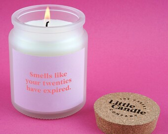May include: A lit white candle in a frosted glass jar with a pink label that reads "Smells like your twenties have expired." The cork lid is next to the jar. The lid has the text "The Cheeky Little Candle Company."