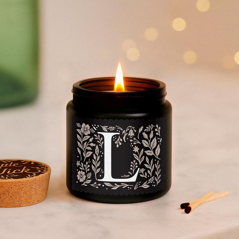 Puede incluir: Una vela negra en un tarro con una mecha encendida. El tarro tiene un dise&ntilde;o floral blanco con la letra "L" en el centro. Una tapa de corcho y cerillas est&aacute;n junto a la vela. Art&iacute;culo decorativo.