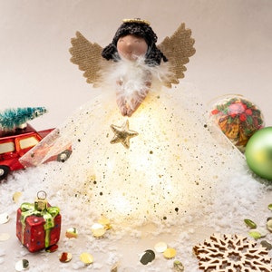 May include: A white angel tree topper with a gold star, surrounded by gold glitter and white snow. The angel has brown hair, a white dress, and gold wings. There are also other Christmas ornaments, including a red car with a small green tree on top, a green glass ornament, and a red gift box.