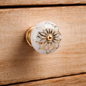 Peut inclure: Un bouton de meuble rond en céramique blanche orné d'un motif floral métallique doré sur la face avant. Le bouton présente une finition lisse et brillante et est fixé sur une base en métal doré reliée à la surface en bois.
