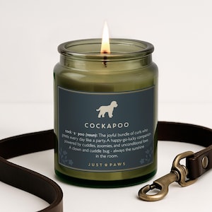 Peut inclure: Une bougie allumée dans un pot en verre vert foncé avec une étiquette présentant une silhouette de chien blanc, le texte COCKAPOO et une définition de la race. L'étiquette inclut le texte JUST PAWS. Une laisse en cuir marron avec un fermoir est présente.