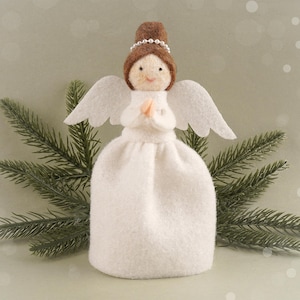 May include: A handcrafted angel figurine with a white dress, wings, and a brown hair bun adorned with a pearl headband. The angel's face is delicately detailed, and its hands are clasped in prayer. The figurine is set against a backdrop of evergreen branches.