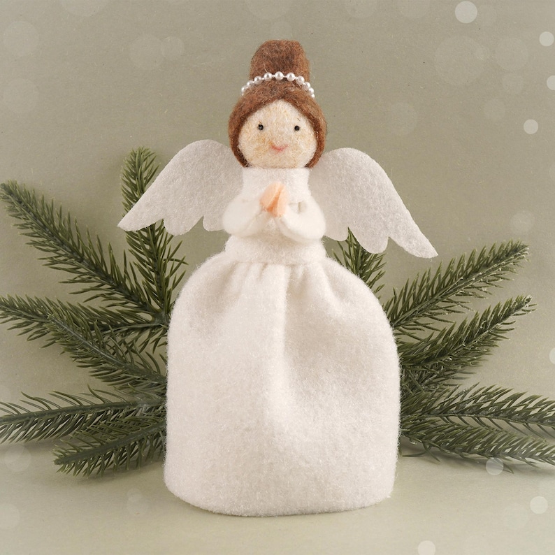 May include: A handcrafted angel figurine with a white dress, wings, and a brown hair bun adorned with a pearl headband. The angel's face is delicately detailed, and its hands are clasped in prayer. The figurine is set against a backdrop of evergreen branches.