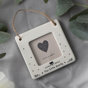 Pode incluir: Moldura de fotos de cerâmica branca com um laço para pendurar. A moldura tem uma pequena abertura para uma foto de 50 mm x 50 mm. A moldura é decorada com corações pretos e o texto "Send with love" e "Our Little Family".