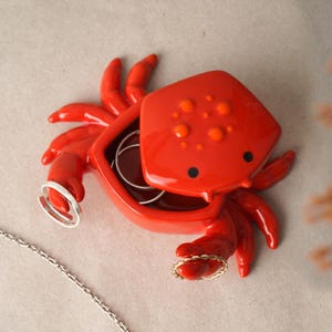 Puede incluir: Un joyero en forma de cangrejo rojo brillante. El plato tiene un acabado brillante, con manchas naranjas en la parte superior y ojos negros. Las pinzas del cangrejo sostienen un anillo plateado y un anillo dorado. El interior del plato tiene una base marrón.
