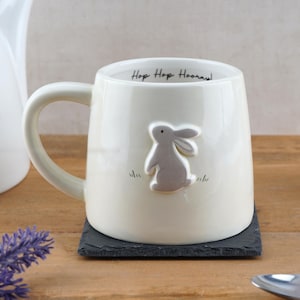 May include: Cream ceramic mug featuring a grey bunny design and the words "Hop Hop Hooray!". The mug has a curved handle and rests on a black square coaster. The mug is on a wooden surface.