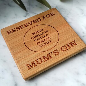 May include: A square wooden coaster with the text "RESERVED FOR MUM'S GIN" and a circular design that reads "WHEN CIRCLE IS VISIBLE, PLEASE REFILL". The coaster is made of light brown wood.
