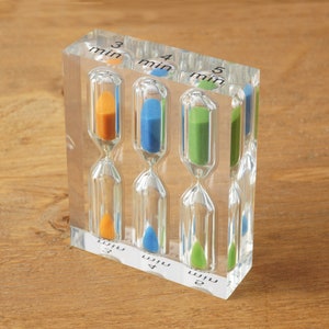 May include: A clear acrylic hourglass timer with four individual sand timers. The timers are labelled 3 min, 4 min, 5 min, and 6 min. The sand is coloured orange, blue, green, and white.